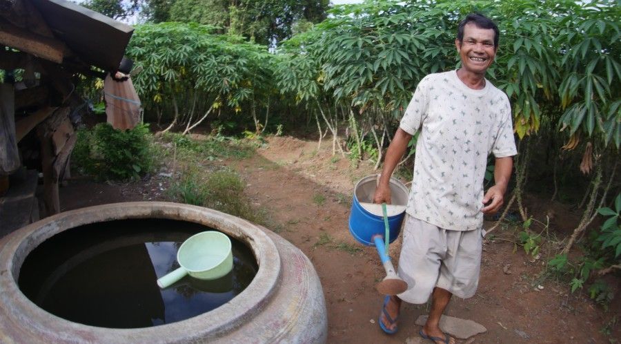 Man standing beside a large round water tank in a garden, holding a blue bucket and smiling