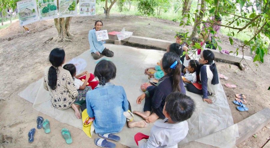 Teacher leading an outdoor lesson to a seated group of children under trees