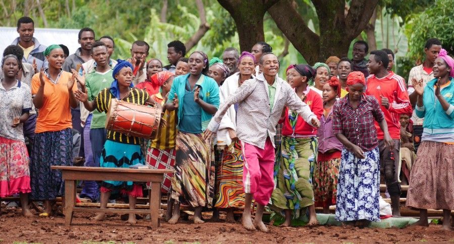 Crowd gathered outdoors around a drum, with people in colorful clothes under trees