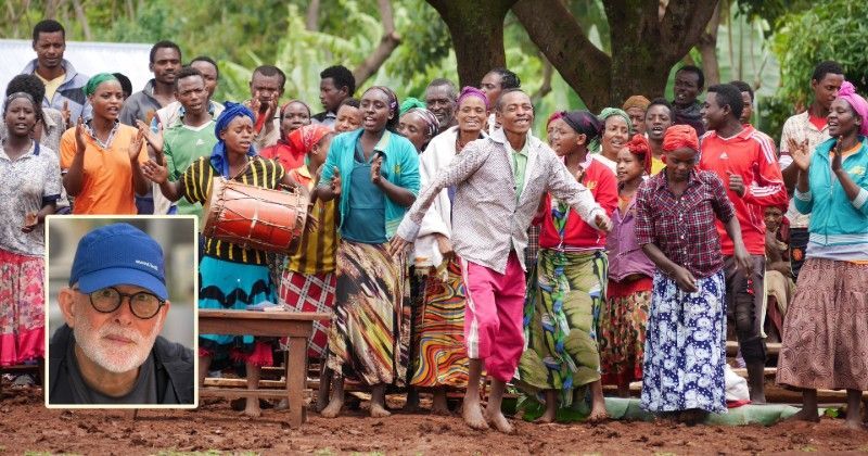 Crowd gathered outdoors around a framed portrait; people wear colorful clothes and hold drums among trees.