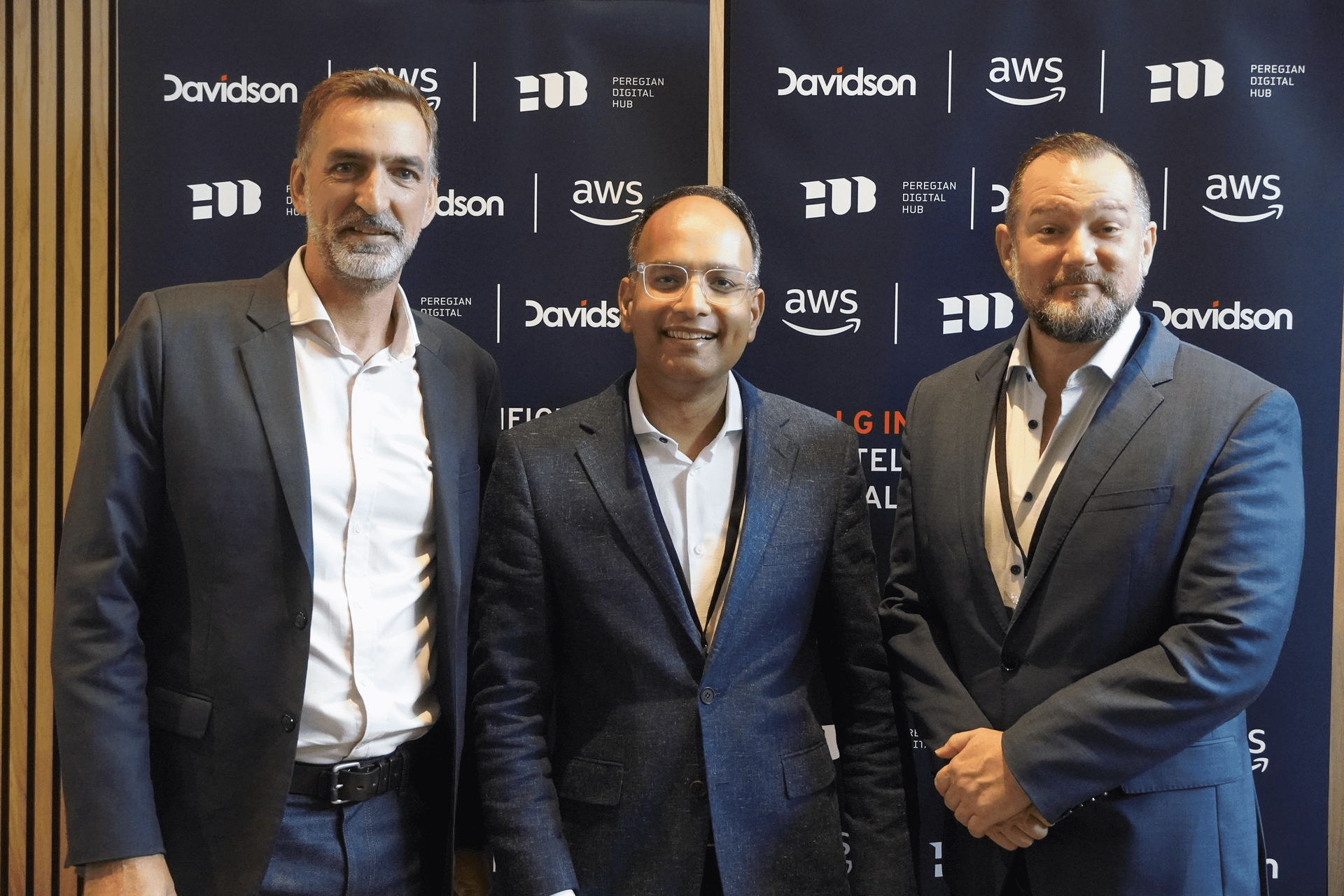Three men in suits are posing for a picture in front of a wall with aws written on it.