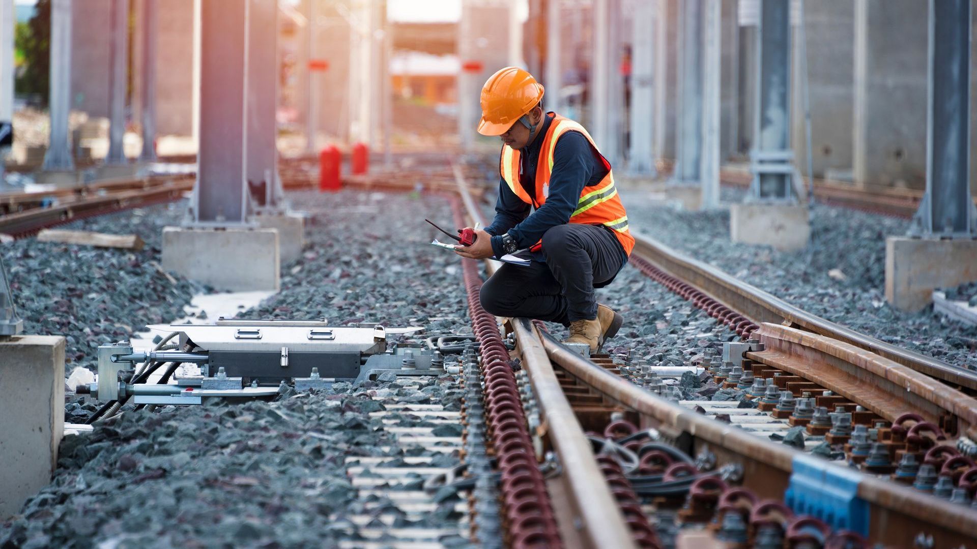 A man in a hard hat and high visibility vest crouches over a railway line and inspects it.