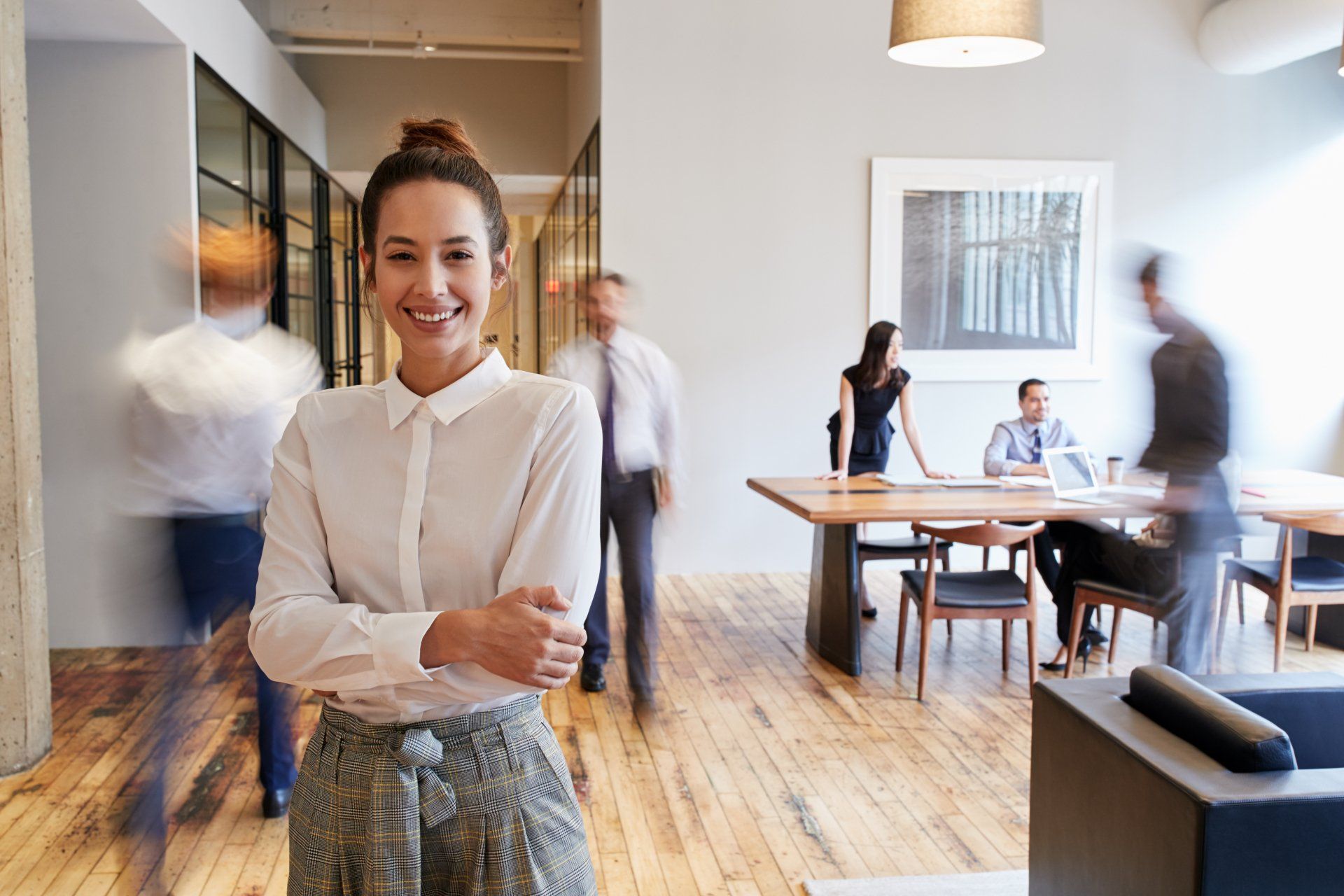 Women standing forward and smiling as her team or coworkers walk behind her or stand around a table.