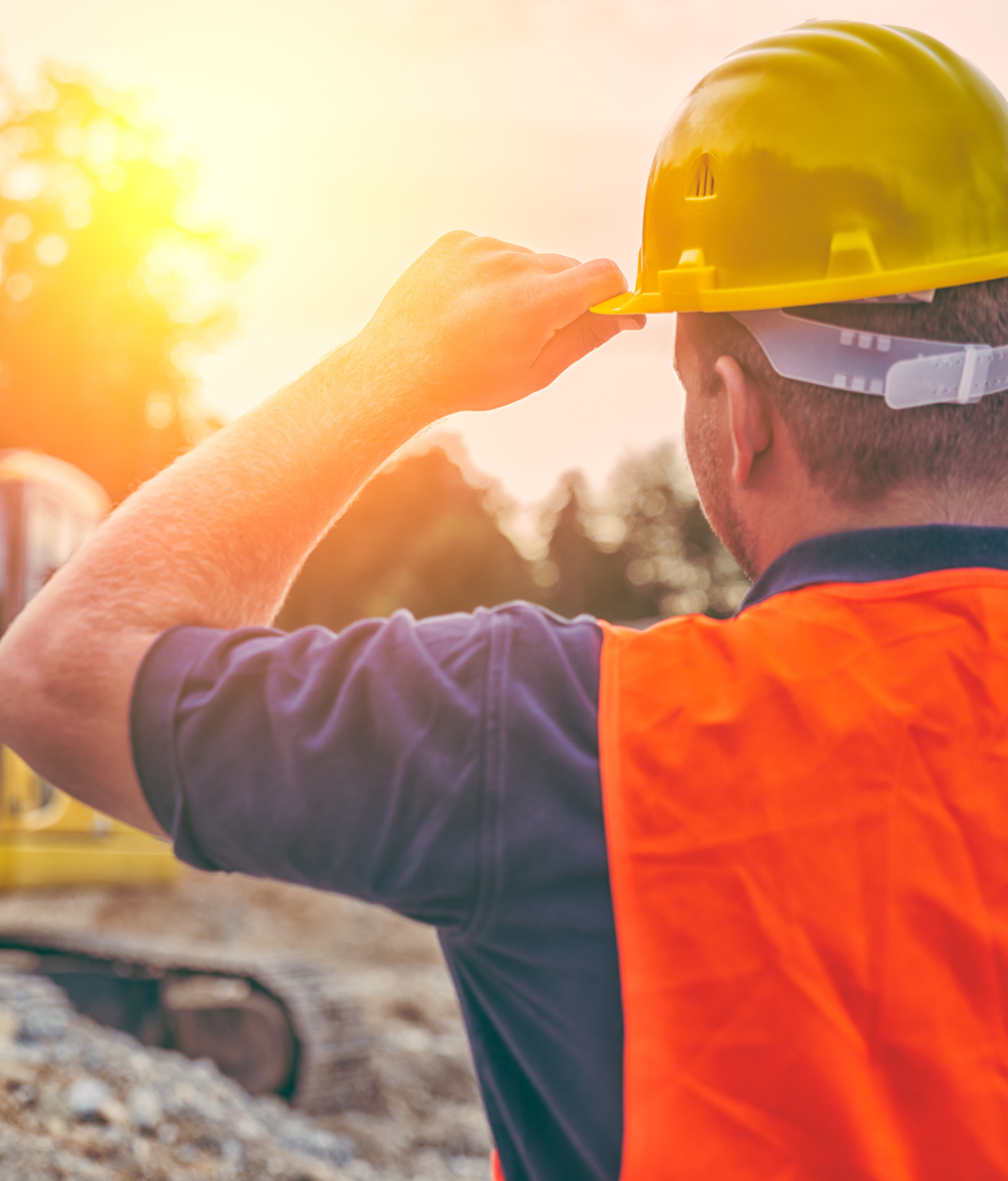 A man looking towards a setting sun, wearing a hard hat and high visibility vest.