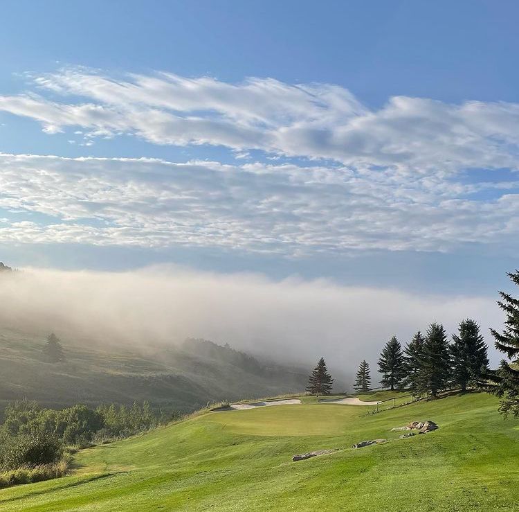 Green golf course with trees, hillside, and low-lying fog under a partly cloudy blue sky.