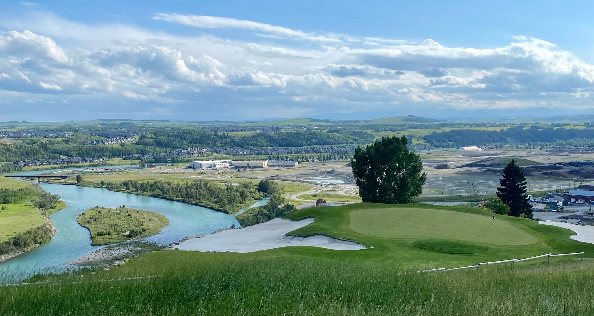 Golf course overlooking a river, town, and cloudy sky. Green grass, sand trap, and trees.