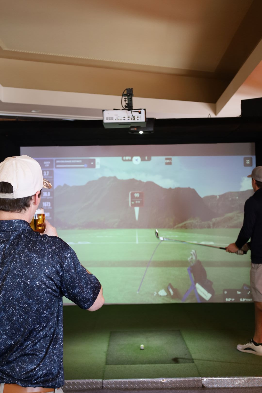 Two men playing golf at an indoor simulator. One holds a beer; the other prepares to swing.