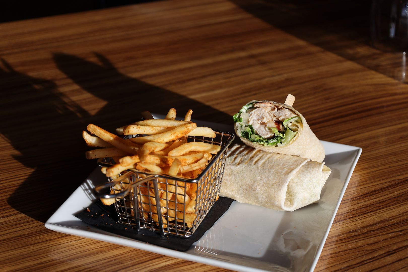 Wrap sandwich with fries in a wire basket on a white square plate, set on a wooden table.