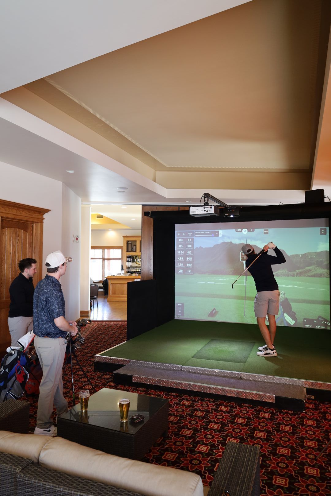 Man swings golf club in simulator room; two others watch.