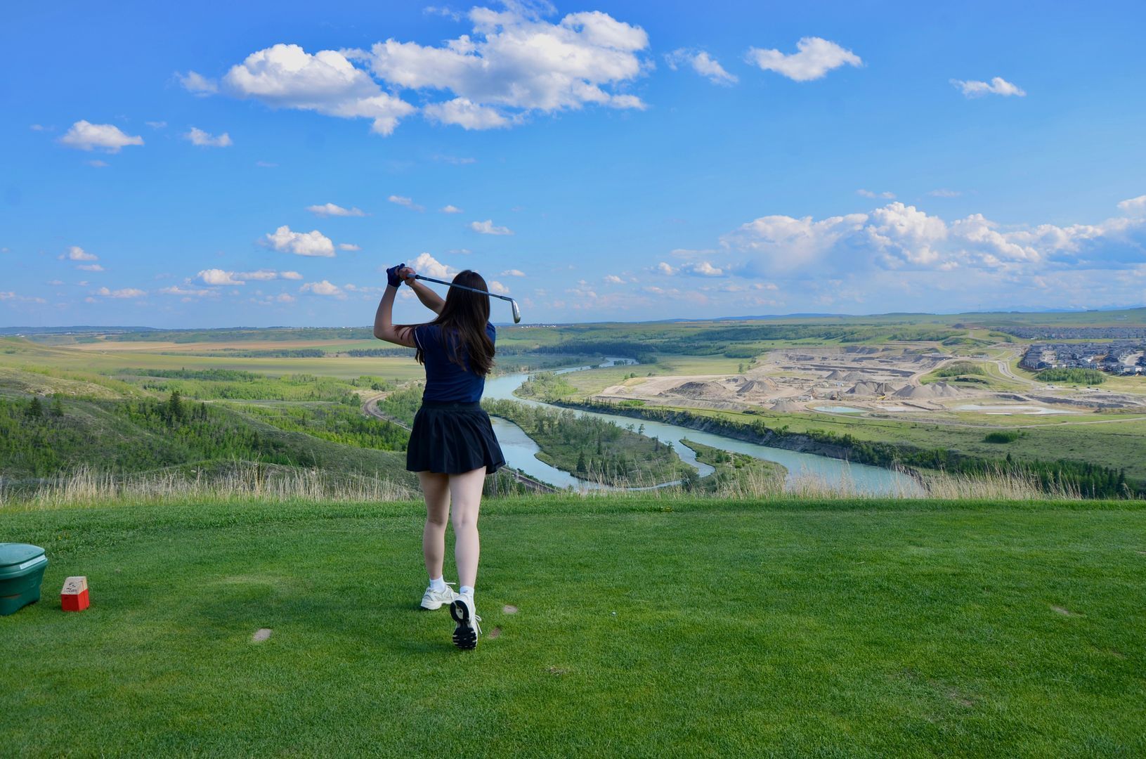 Person swinging a golf club on a green, scenic view of valley and river under a blue sky.