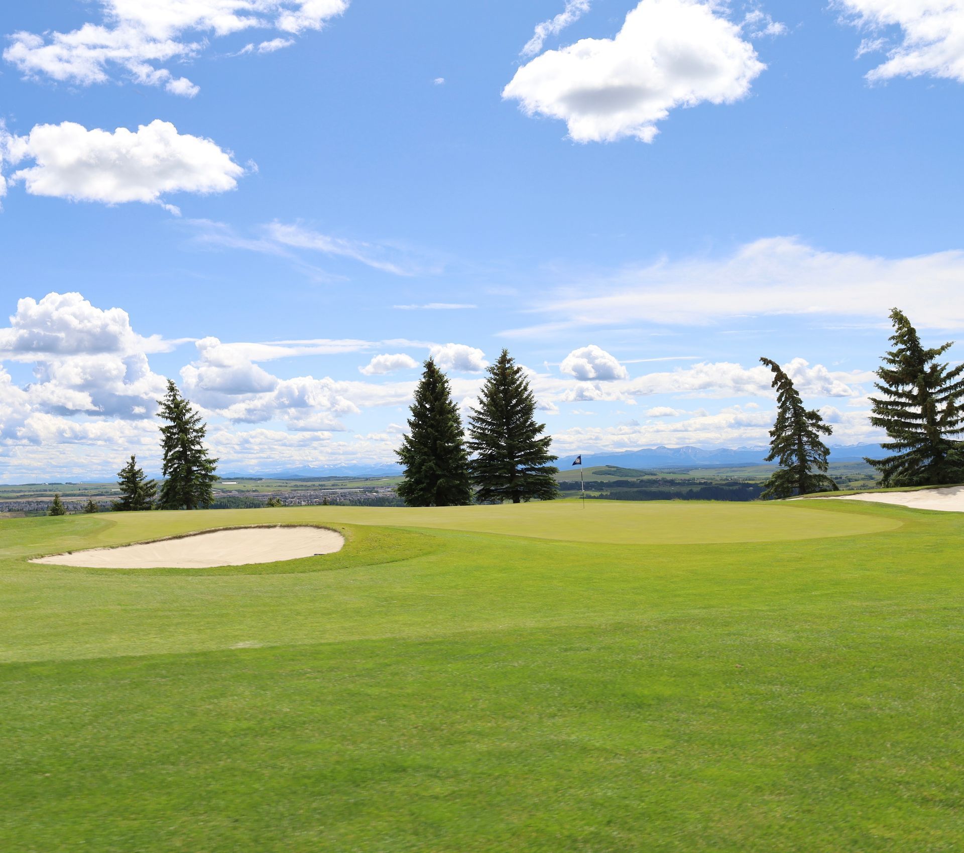 Green golf course with sand trap and trees under a blue sky with clouds.