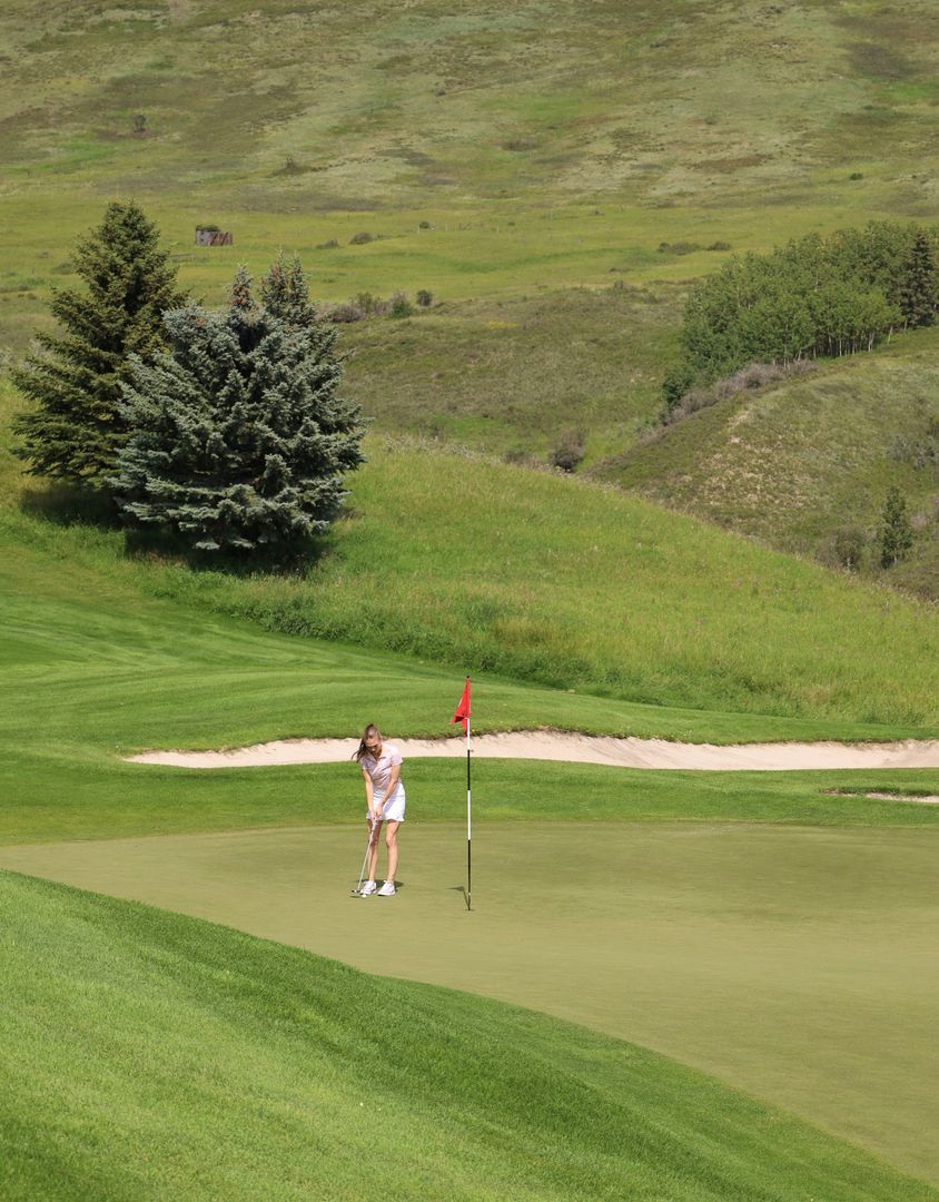 Golfer about to putt on a green. Green grass, sand trap, red flagstick, hillside in the background.