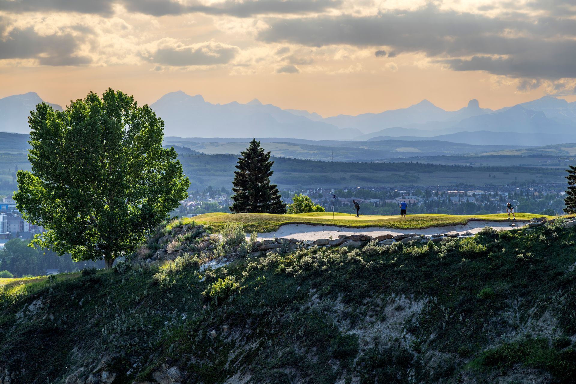 Golfers on a green course, blue golf cart, sunny day. Homes and grassy hill in background.