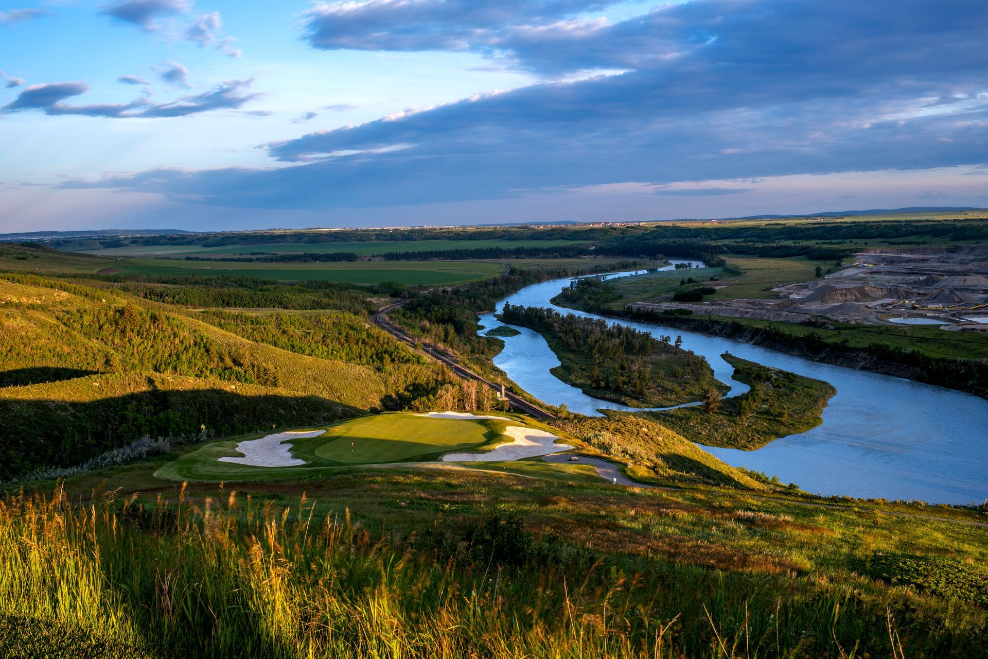 Green golf course overlooking a river and hills under a clear blue sky.