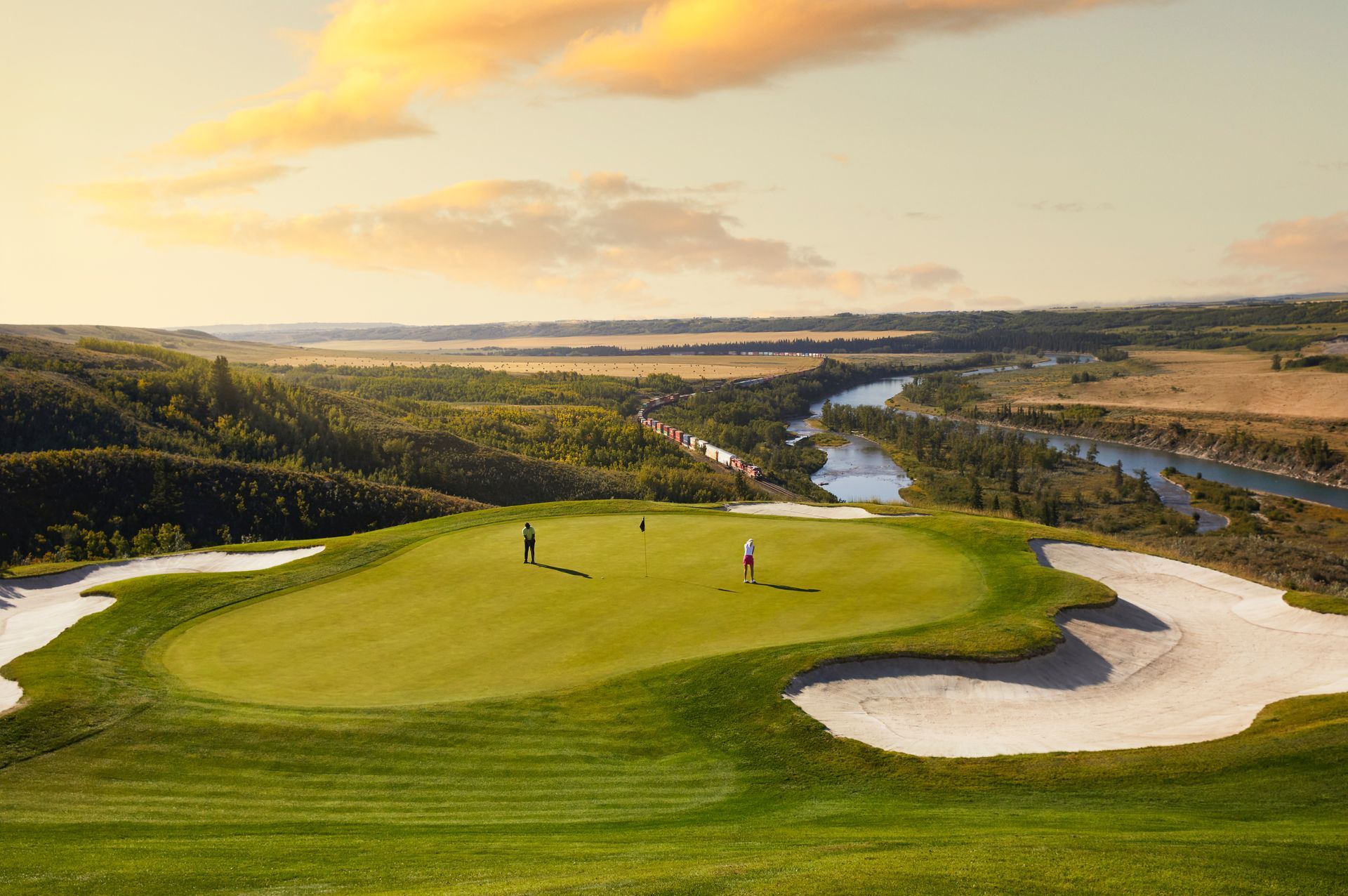 Golfers putting on a green, set against a valley, with mountains in the background.