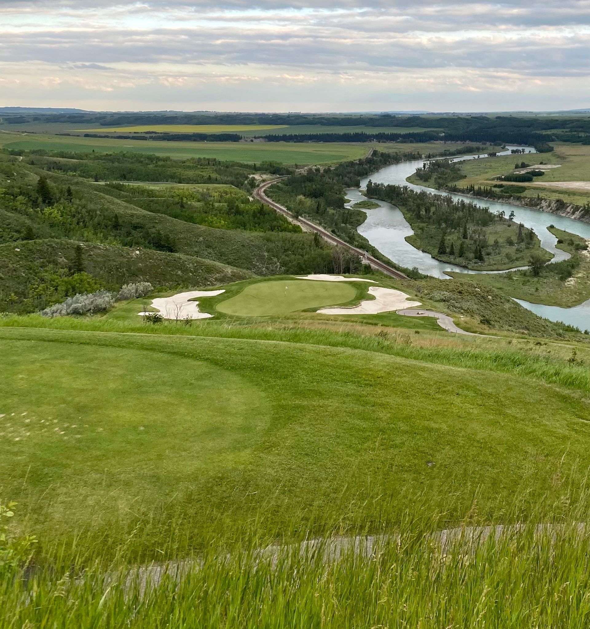 Golf course overlooking a river, green fairways and sand traps. Cloudy sky.
