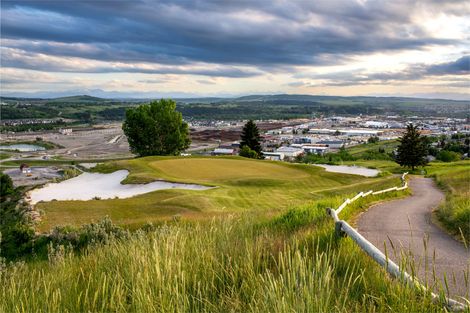 Aerial view of a green golf course with a paved path, bunkers, and trees. City skyline in the background.