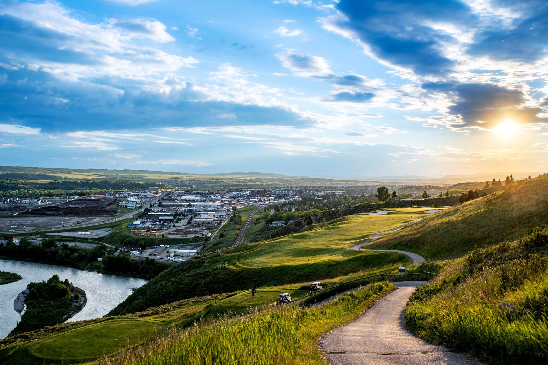 Rolling green hills overlook a city and river under a bright, partly cloudy sky; sun shines.