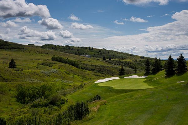 Green golf course with a sand trap, green grass, and hills under a blue, cloudy sky.