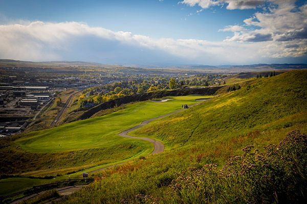 Green golf course on a hillside overlooking a town, with a cloudy blue sky above.