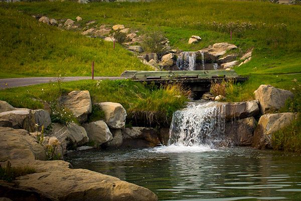 Small waterfalls cascade into a pond, surrounded by rocks and green grass.