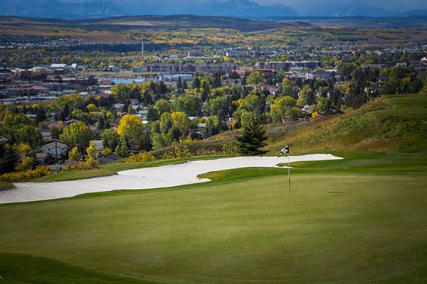Green golf course overlooking a city with mountains in the distance; a bunker is in the foreground.