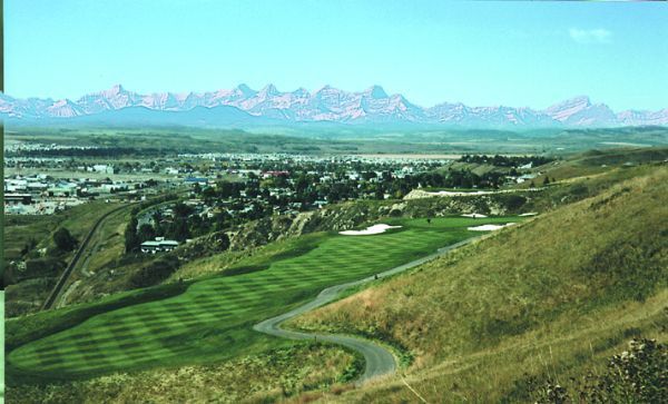 Green golf course, town, and mountains under blue sky.
