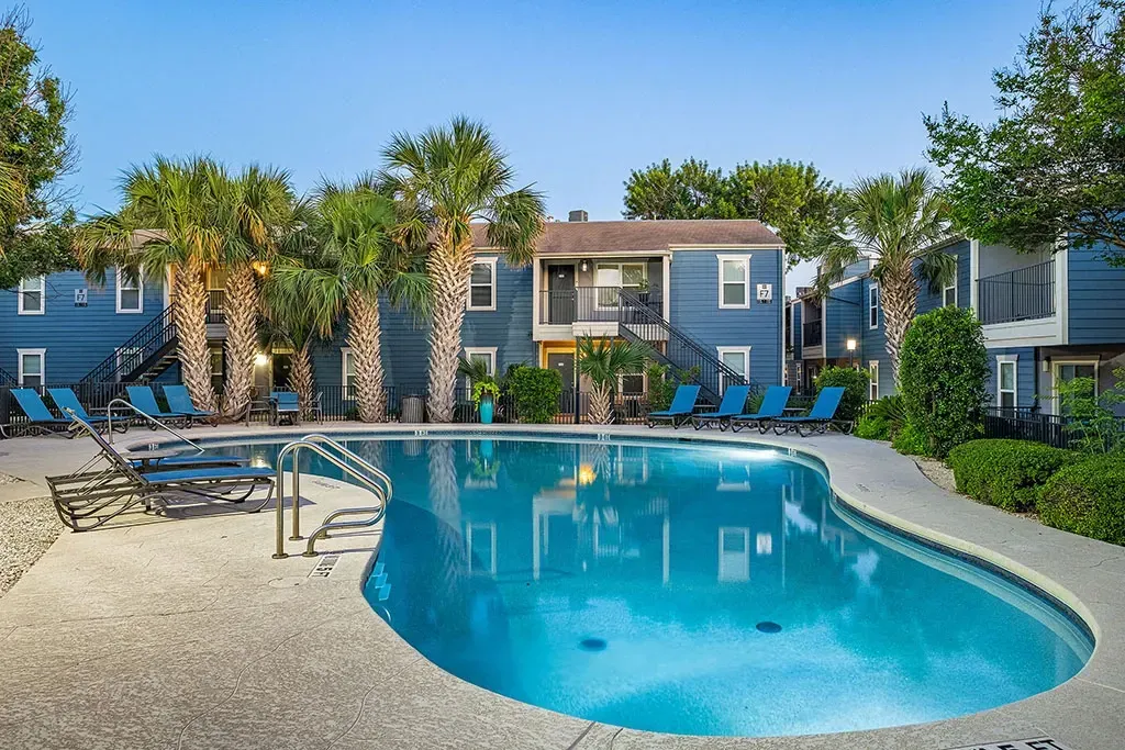 Blue swimming pool with lounge chairs and palm trees in front of blue apartment buildings.