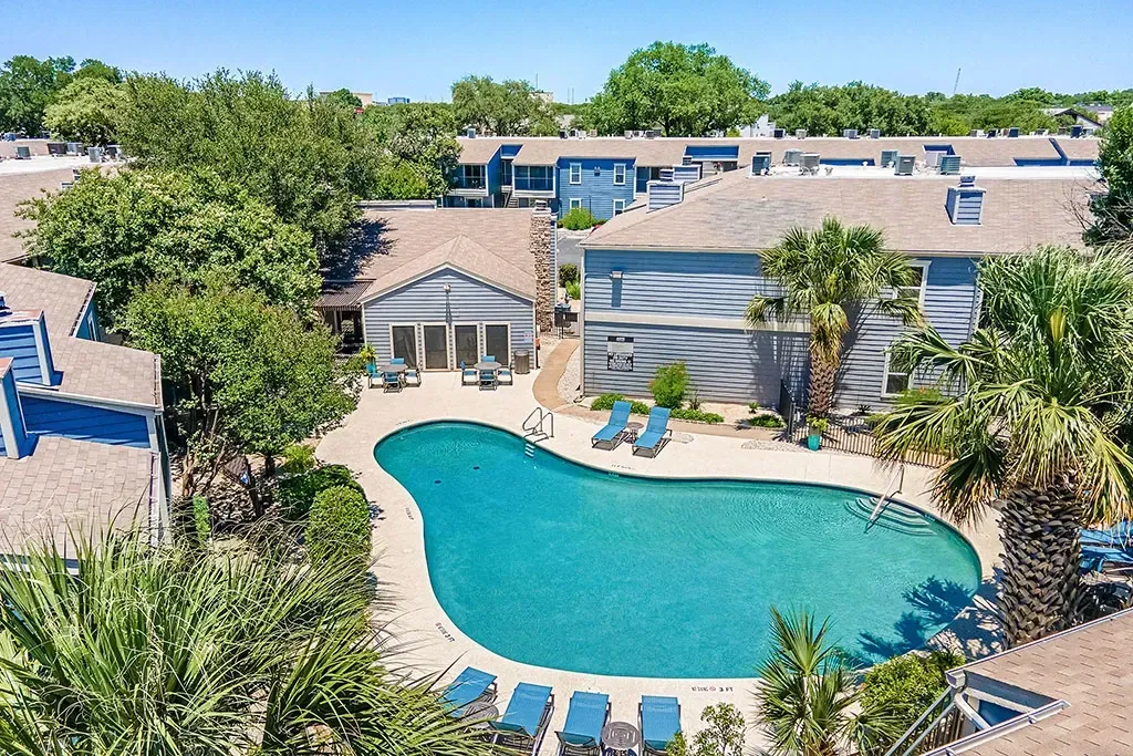 Aerial view of a shimmering blue swimming pool surrounded by lounge chairs and palm trees, with apartment buildings in the background.