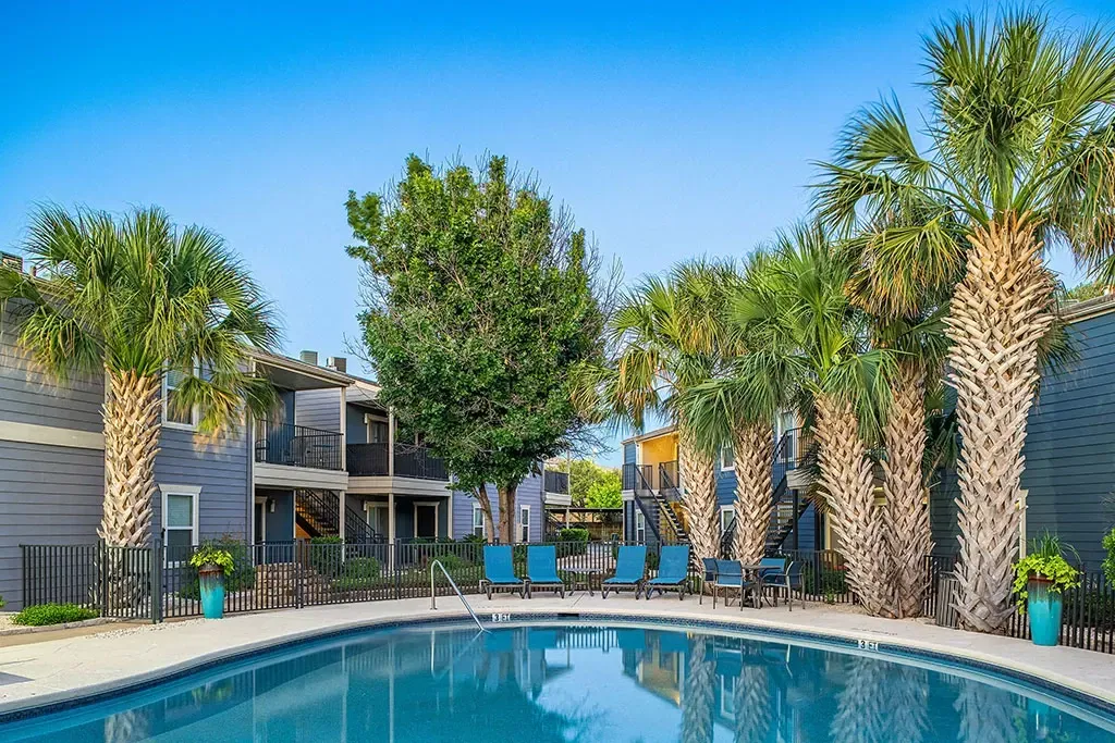 Outdoor swimming pool with lounge chairs and palm trees, apartment buildings in the background.