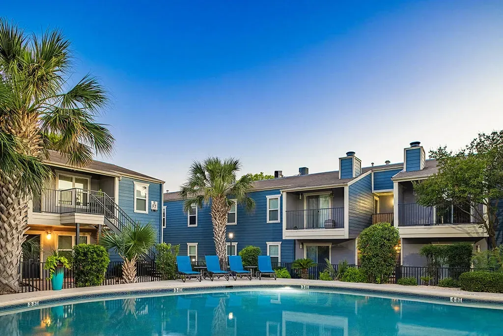 Blue apartment buildings with balconies surround a sparkling swimming pool with lounge chairs.