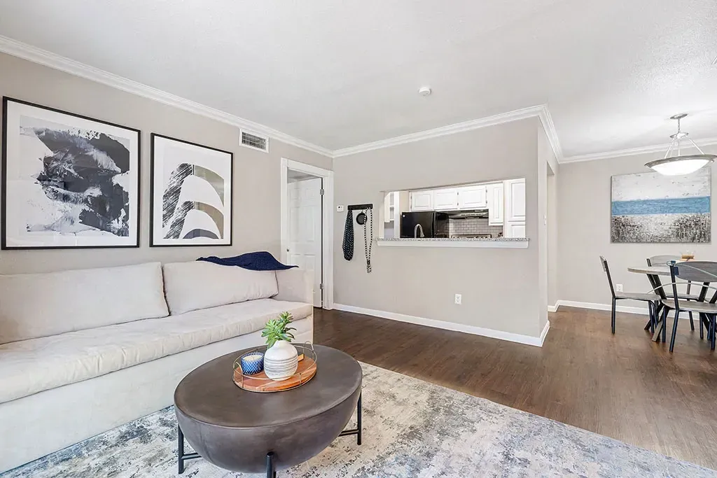 Modern living room with a large sofa, coffee table, and abstract art, opening to a kitchen.
