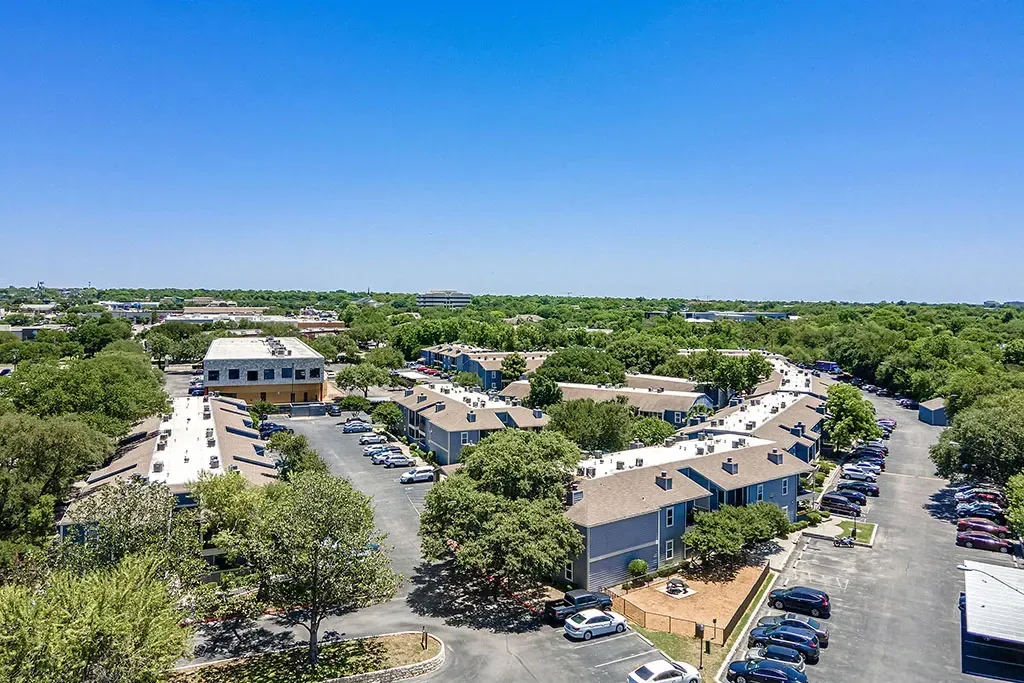Aerial view of apartment buildings with surrounding trees and parking lots under a clear blue sky.