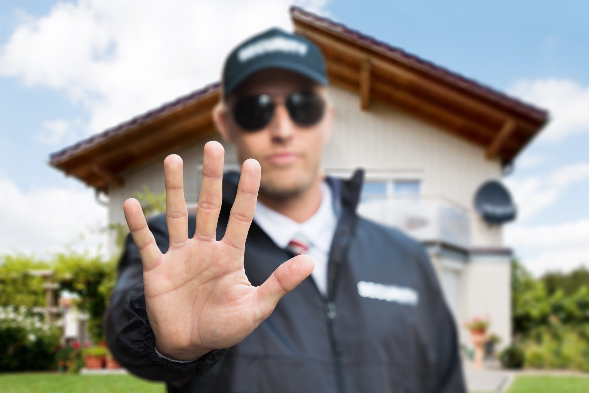 A man wearing sunglasses and a hat is making a stop sign with his hand in front of a house.