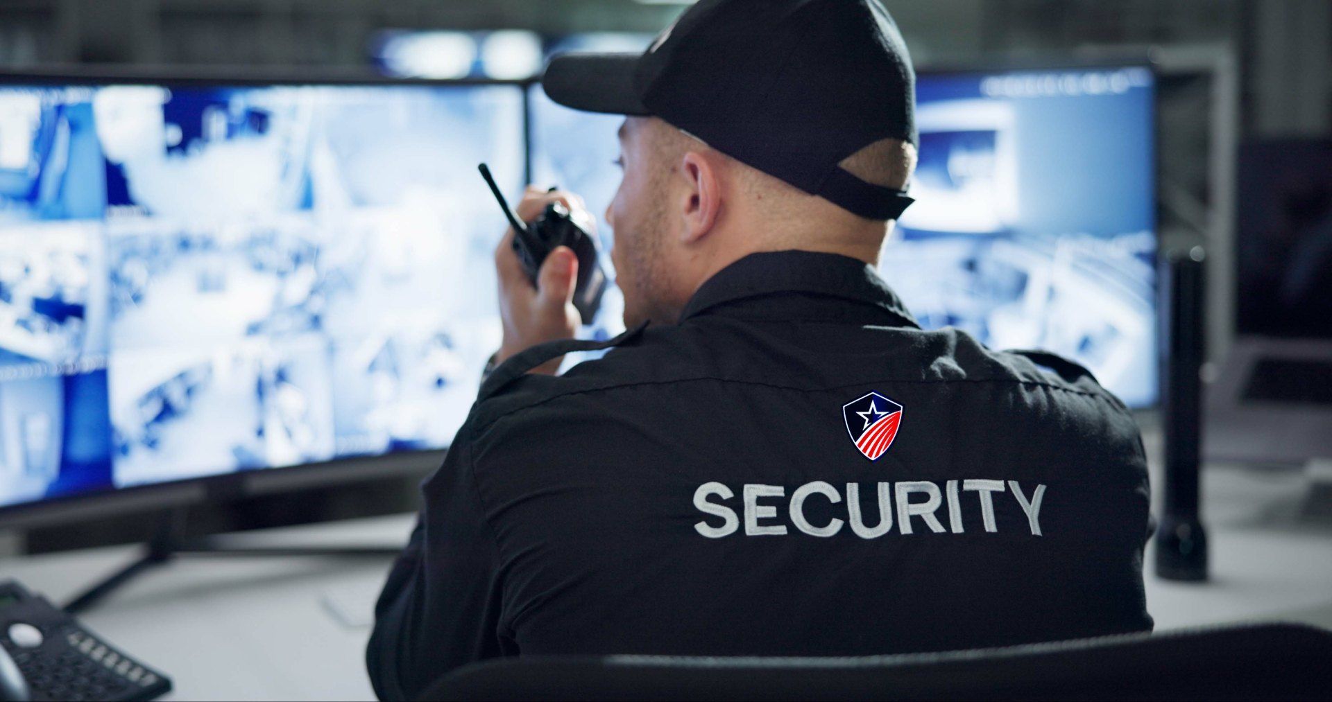 A security guard is sitting at a desk talking on a walkie talkie.