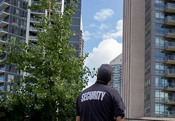 A man wearing a security shirt is standing in front of a tall building.
