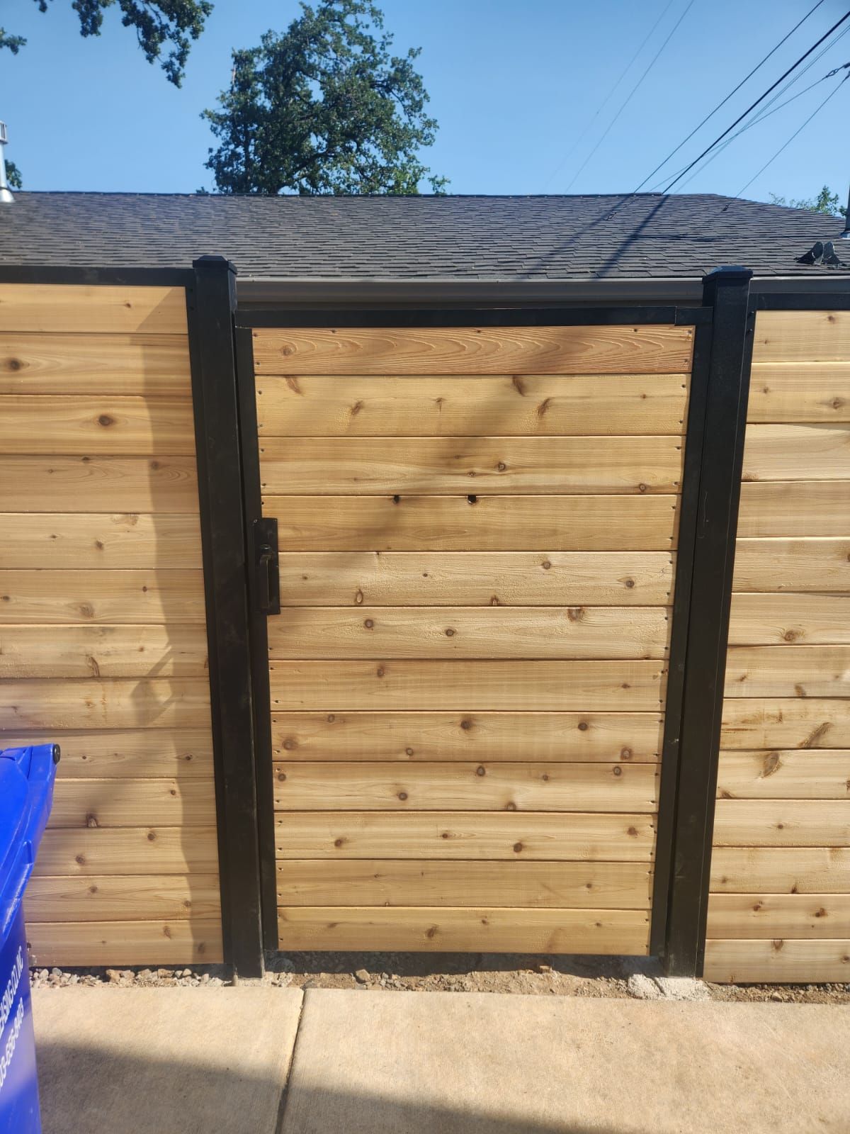 A wooden fence with a black gate and a blue trash can in front of it