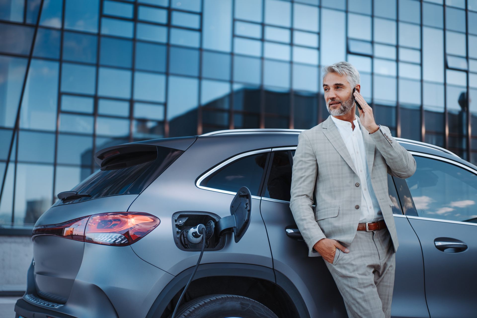 A man in a suit is leaning against a car while talking on a cell phone.
