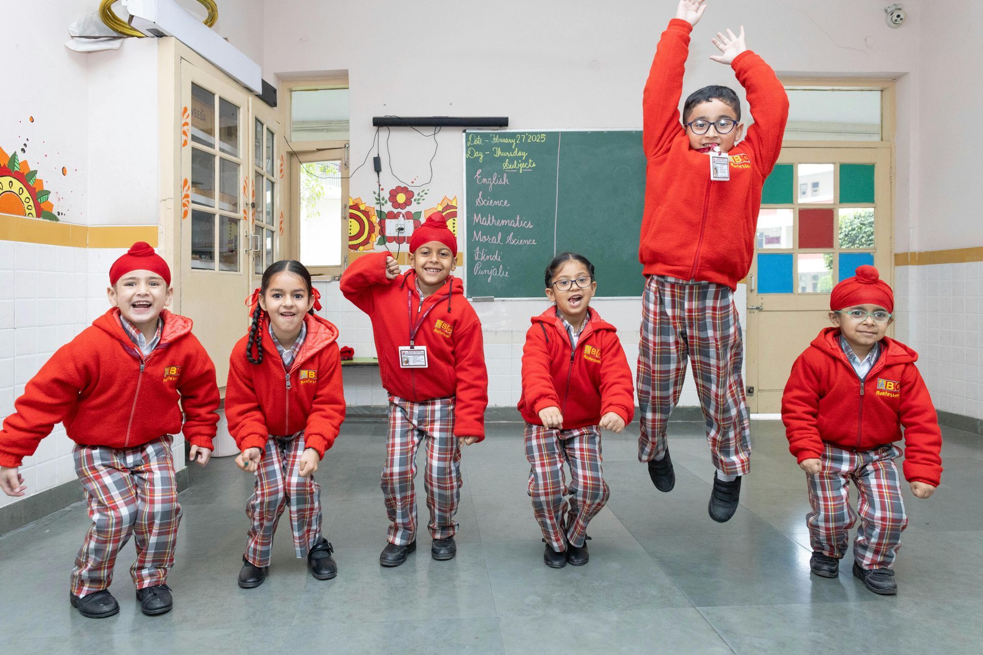 Children in red jackets and plaid pants, jumping and smiling in a classroom.