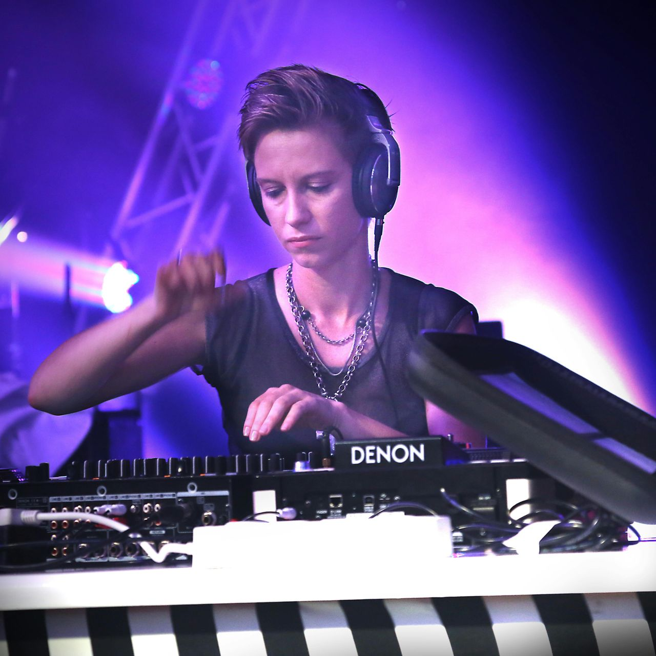 A person wearing headphones DJs on a Denon console under purple stage lights.
