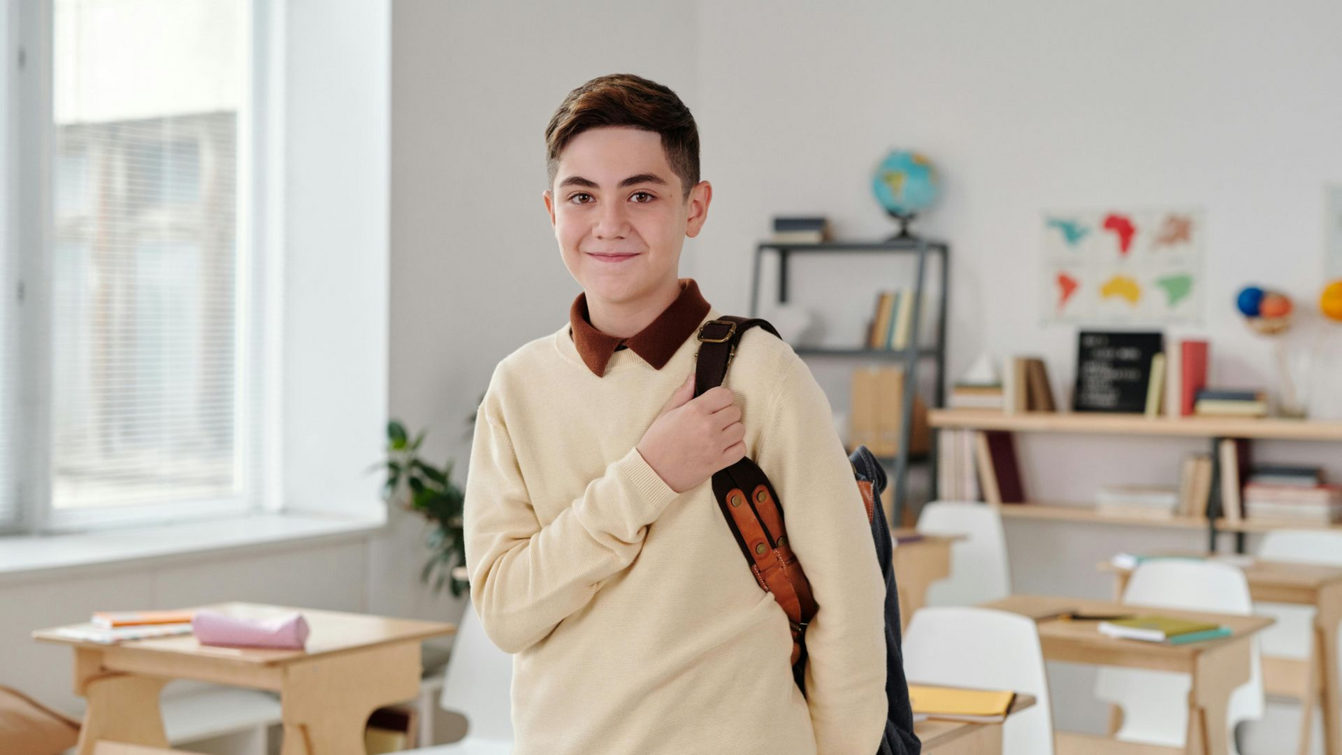 Teenager with backpack smiling in a classroom, standing near a desk.