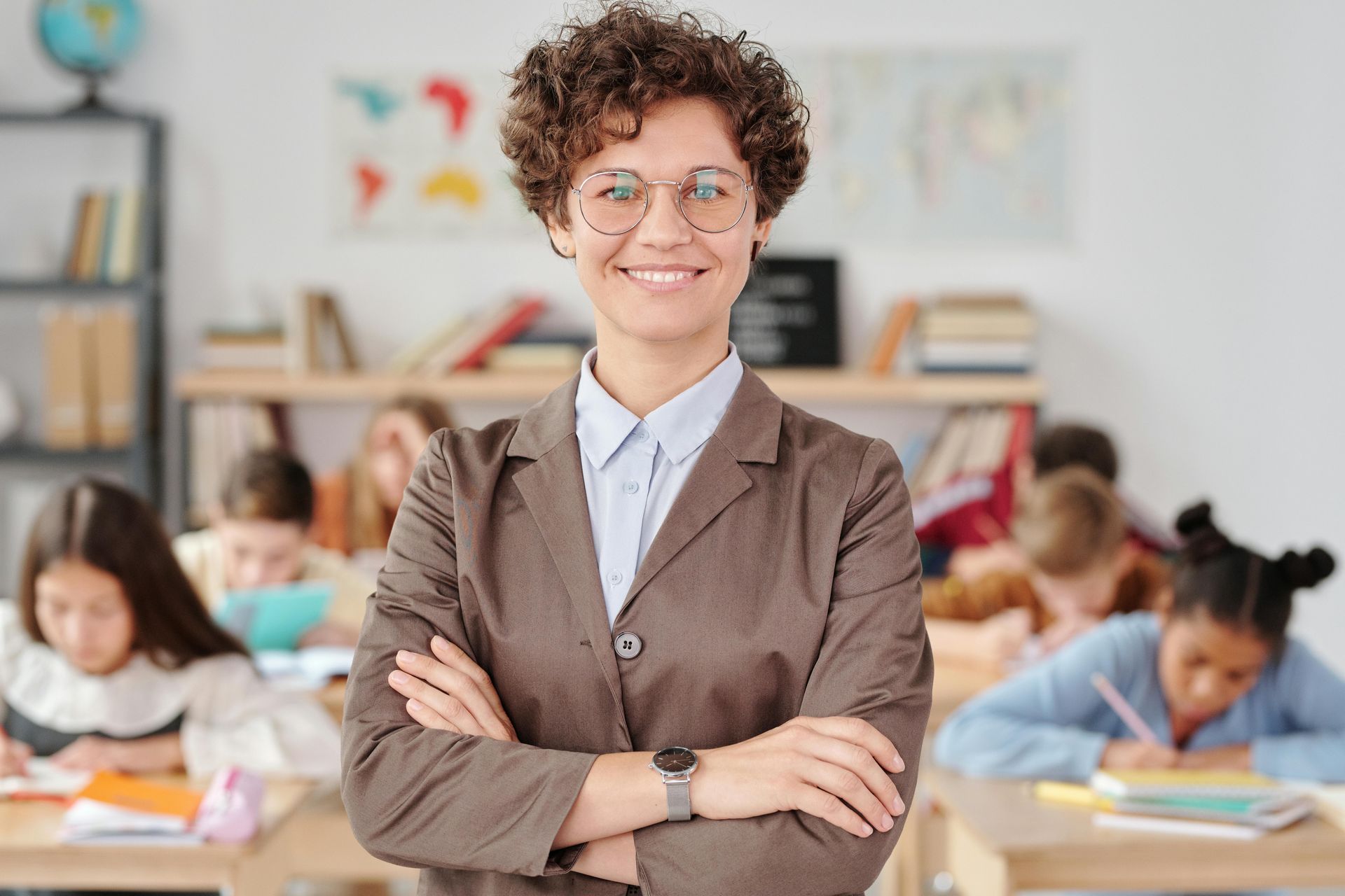 Teacher with arms crossed smiling in a classroom with students working at desks.