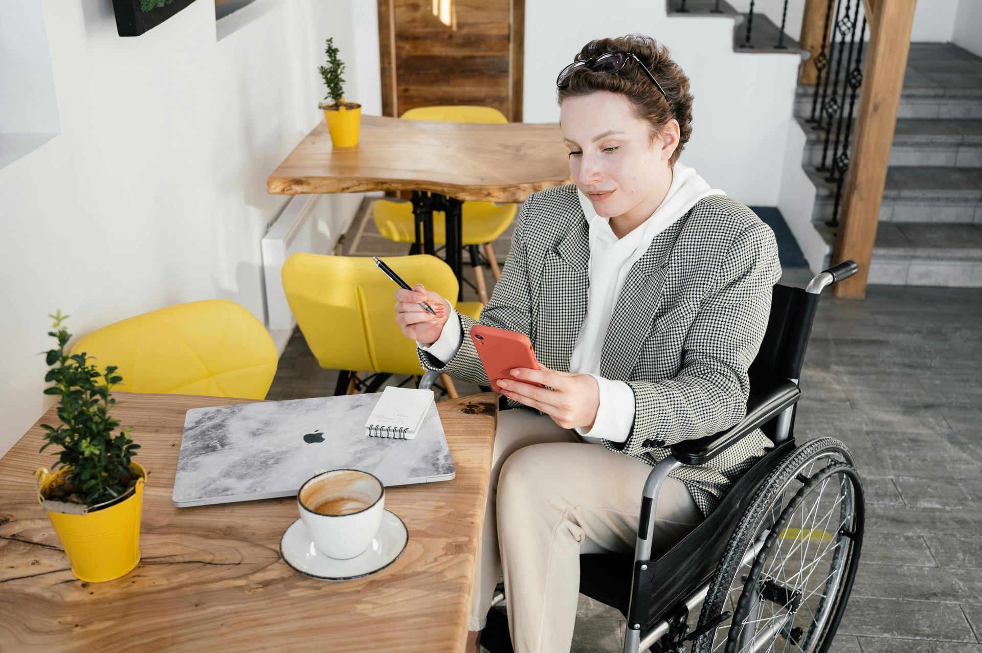 Woman in wheelchair using phone, holding pen, at cafe table with laptop, coffee, and plants.