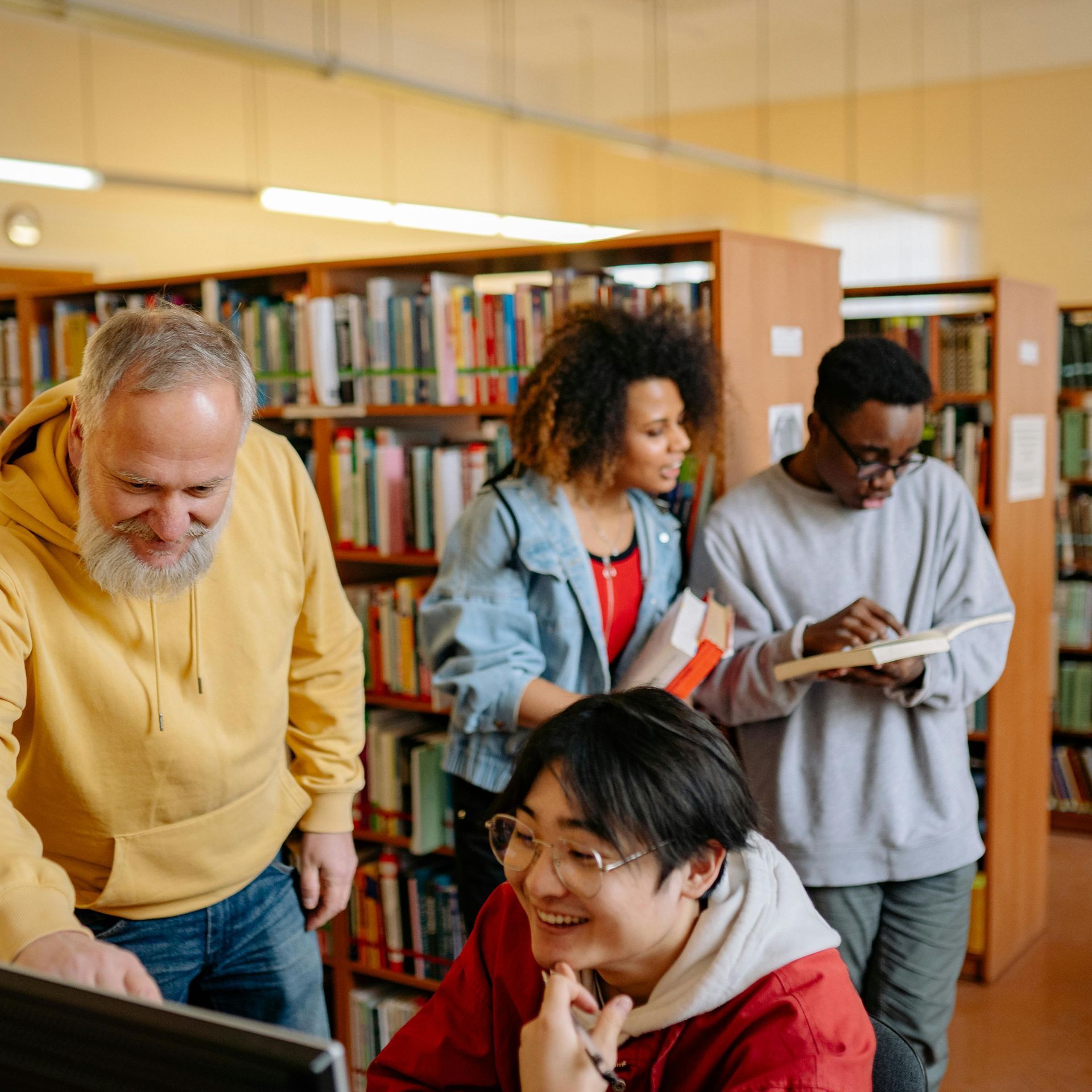 People in a library: looking at a computer, holding books, and taking notes near bookshelves.