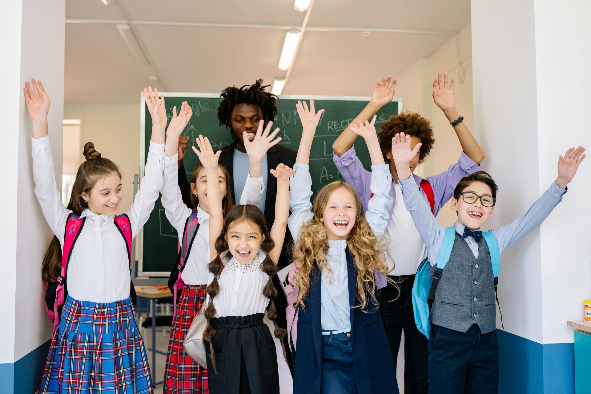 A teacher and students raise arms in a school hallway with a chalkboard.