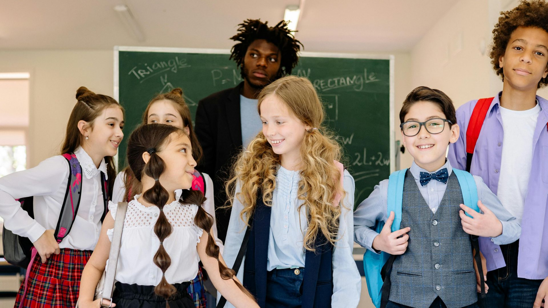 Students and teacher smiling in a classroom with a chalkboard. Several wear backpacks.