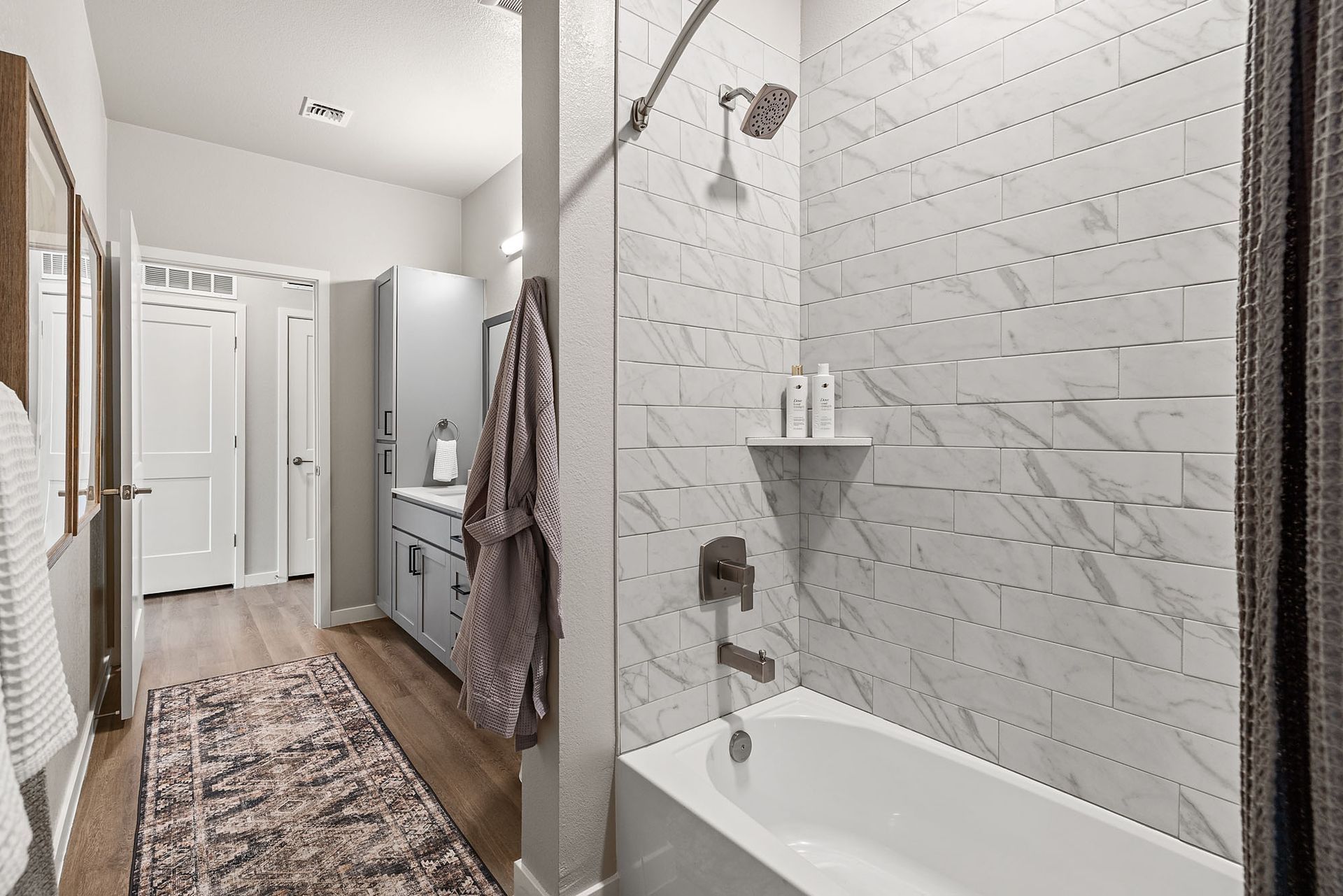 Bathroom with white tile shower, bathtub, vanity, and wood-look flooring at Marq Bronco Butte, offers 1, 2, and 3 bedroom apartments in North Phoenix, AZ. A rug and robe hang inside.