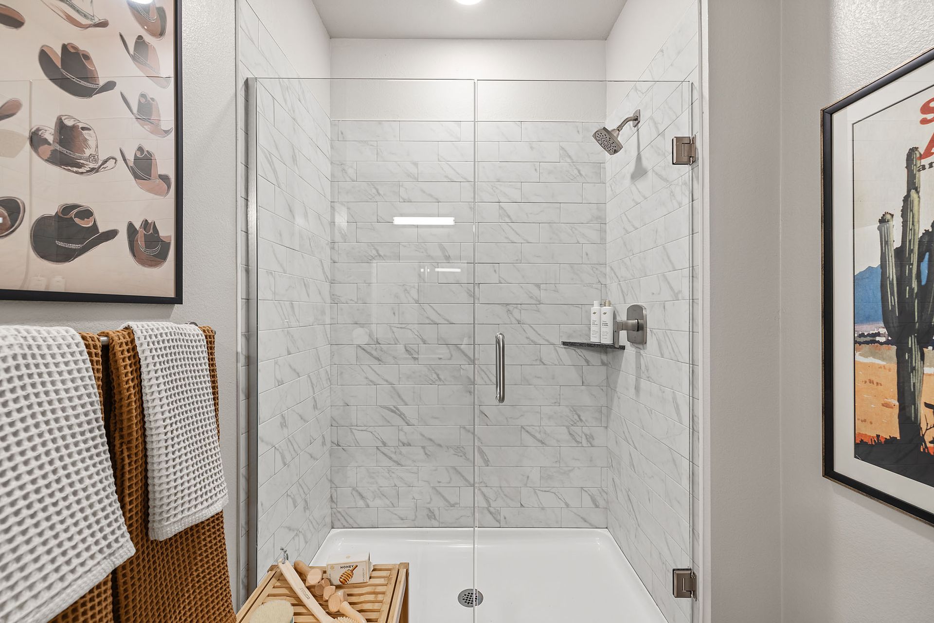 Shower with glass door, marble-like tile walls, and showerhead at Marq Bronco Butte, offers loft-style apartments in North Phoenix, AZ. Towel rack and art on the walls.