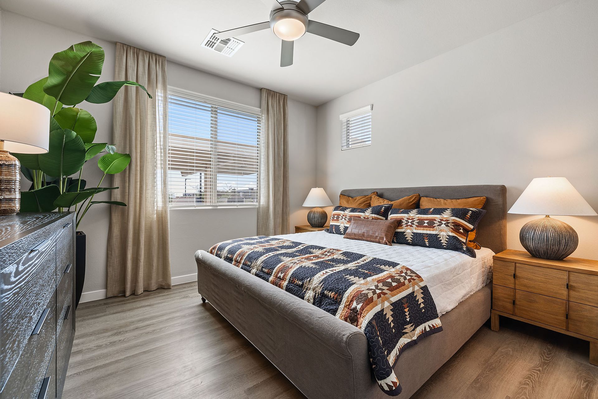 Bedroom with bed, nightstands, window, dresser, and ceiling fan at Marq Bronco Butte, offers loft-style apartments in North Phoenix, AZ. Gray, beige, brown tones.