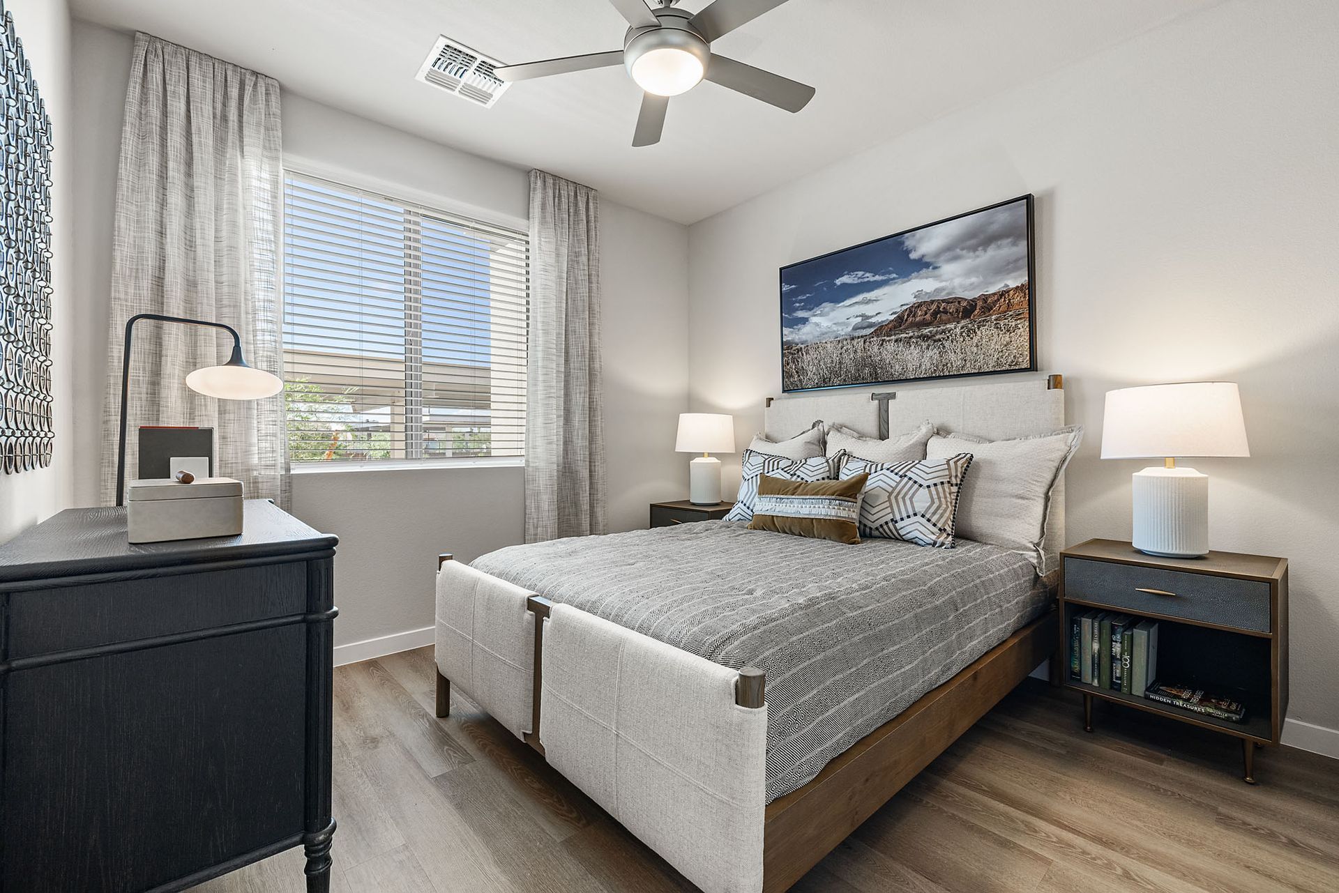 Bedroom with bed, nightstands, window, dresser, artwork, ceiling fan, and light-colored wood flooring at Marq Bronco Butte, offers loft-style apartments in North Phoenix, AZ.